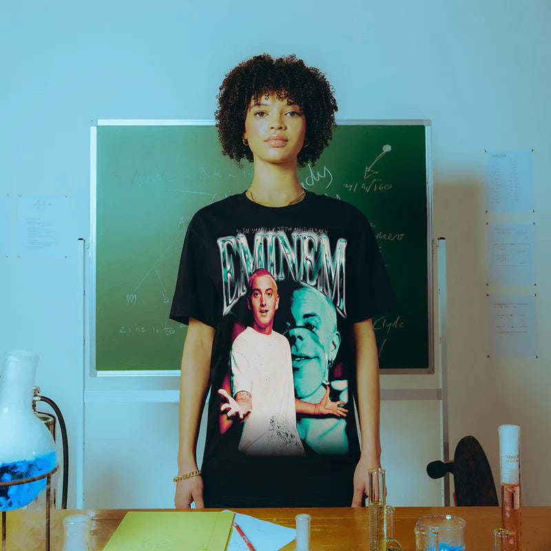 Model wearing the Eminem SSLP25 Chrome T-shirt in a classroom setting, displaying a vibrant print of Eminem's early persona with chrome-style lettering.