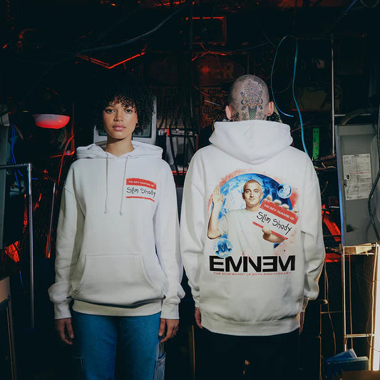 A male and female model facing opposite directions, showing off the front 'Hi My Name Is' name tag and the back graphic with Eminem's portrait on the SSLP25 Name Tag Hoodie.